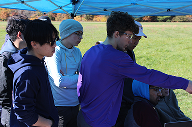 students standing outside under a tent looking at a screen thats out of view 