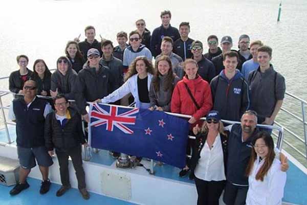 group of dozen-plus students at helm of boat holding New Zealand flag