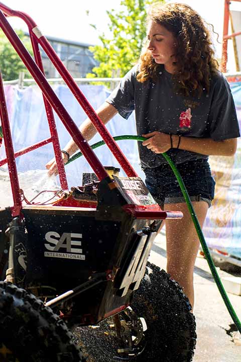 student with hose spraying water to wash the Baja vehicle outside on sunny day